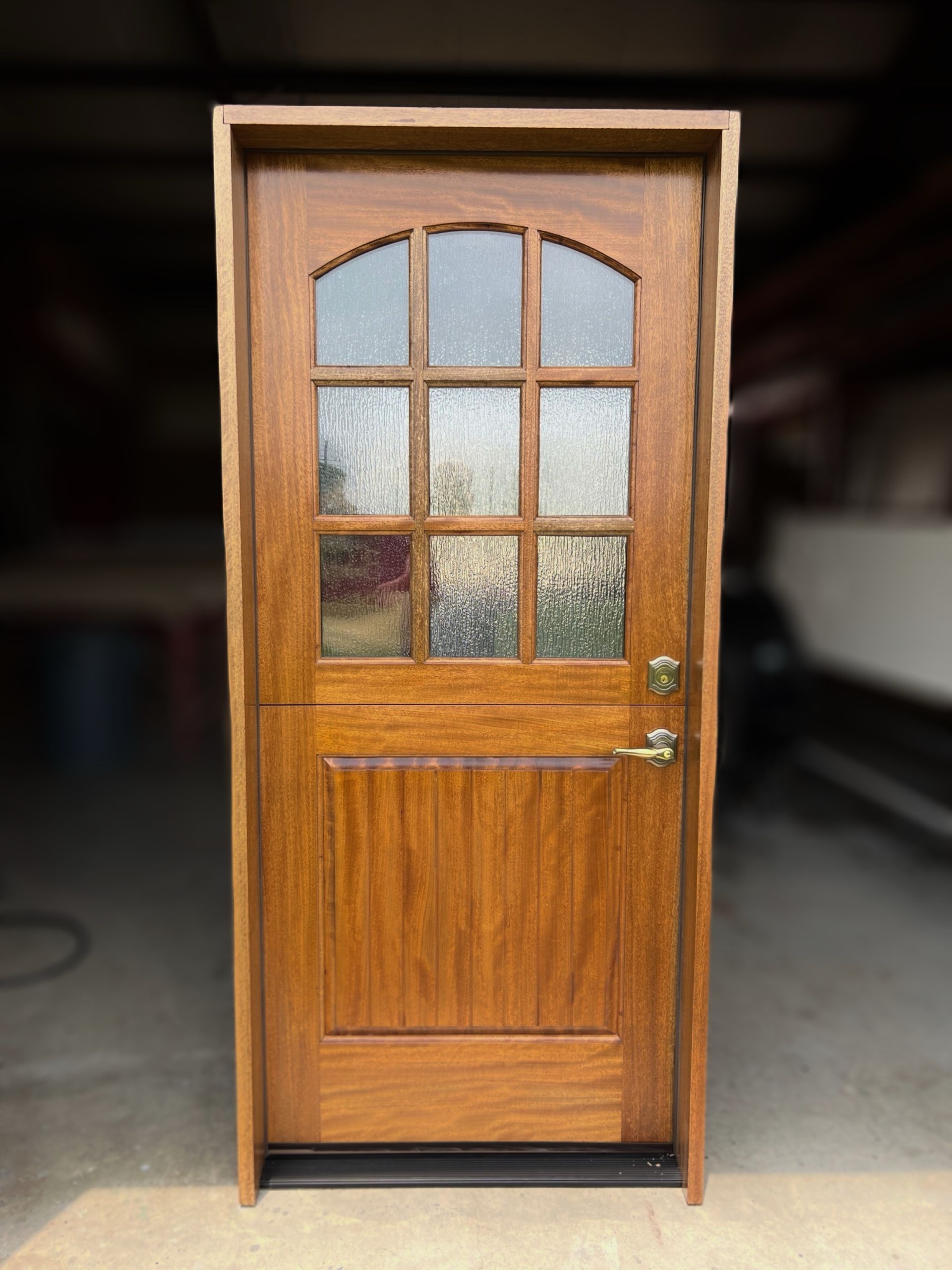 Wooden front door with arched window panes and brass handle, standing in a garage or workshop.
