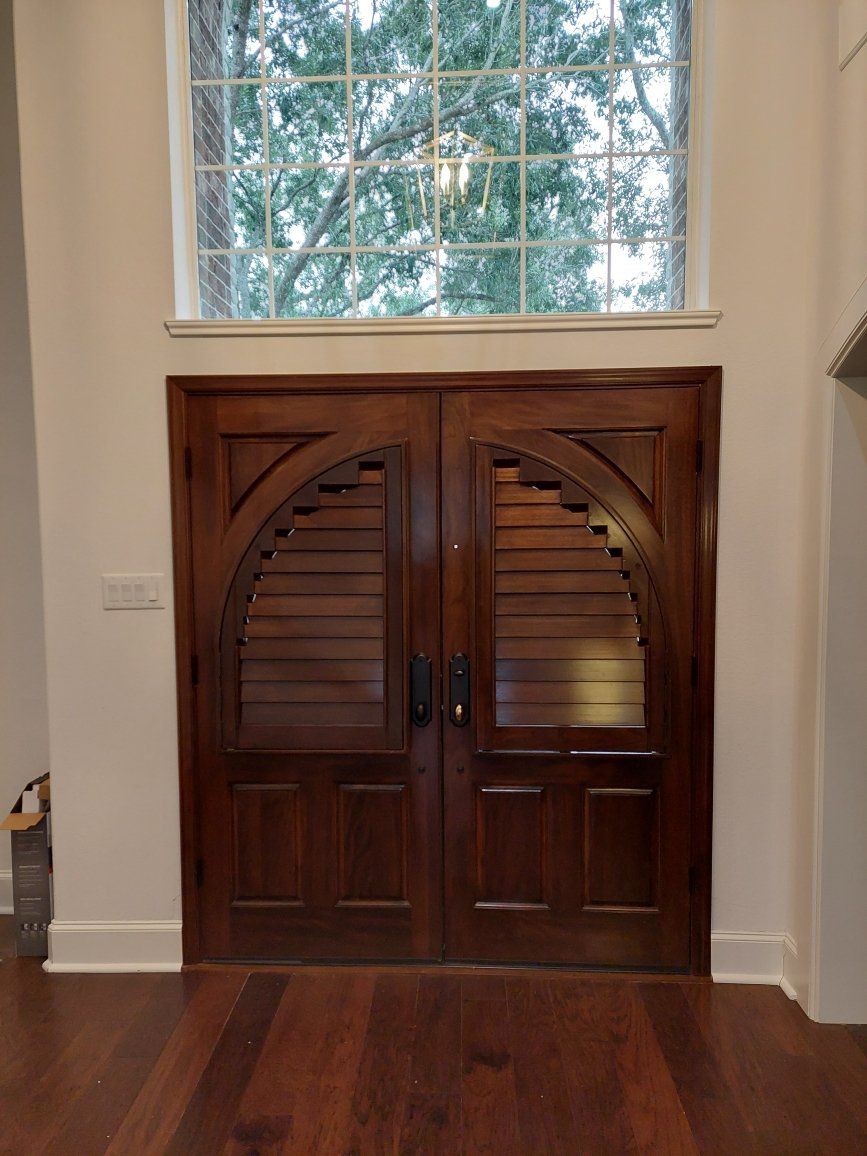 Wooden double doors with arched glass panels beneath a large window in a hardwood-floored room