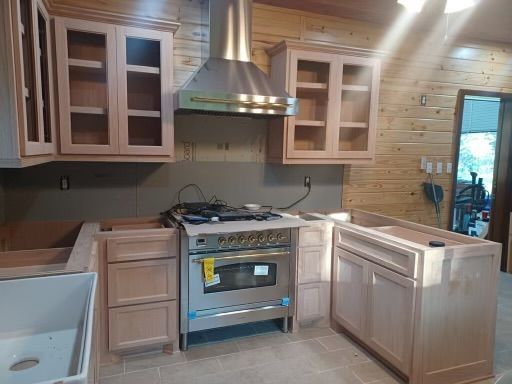 Unfinished kitchen with light wood cabinets, stainless stove, sink, and gray backsplash beside a window.
