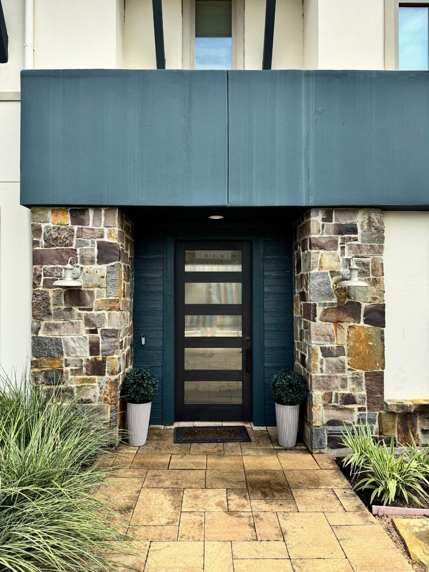 Modern front door with dark blue awning, stone facade, and potted plants along a paved entryway