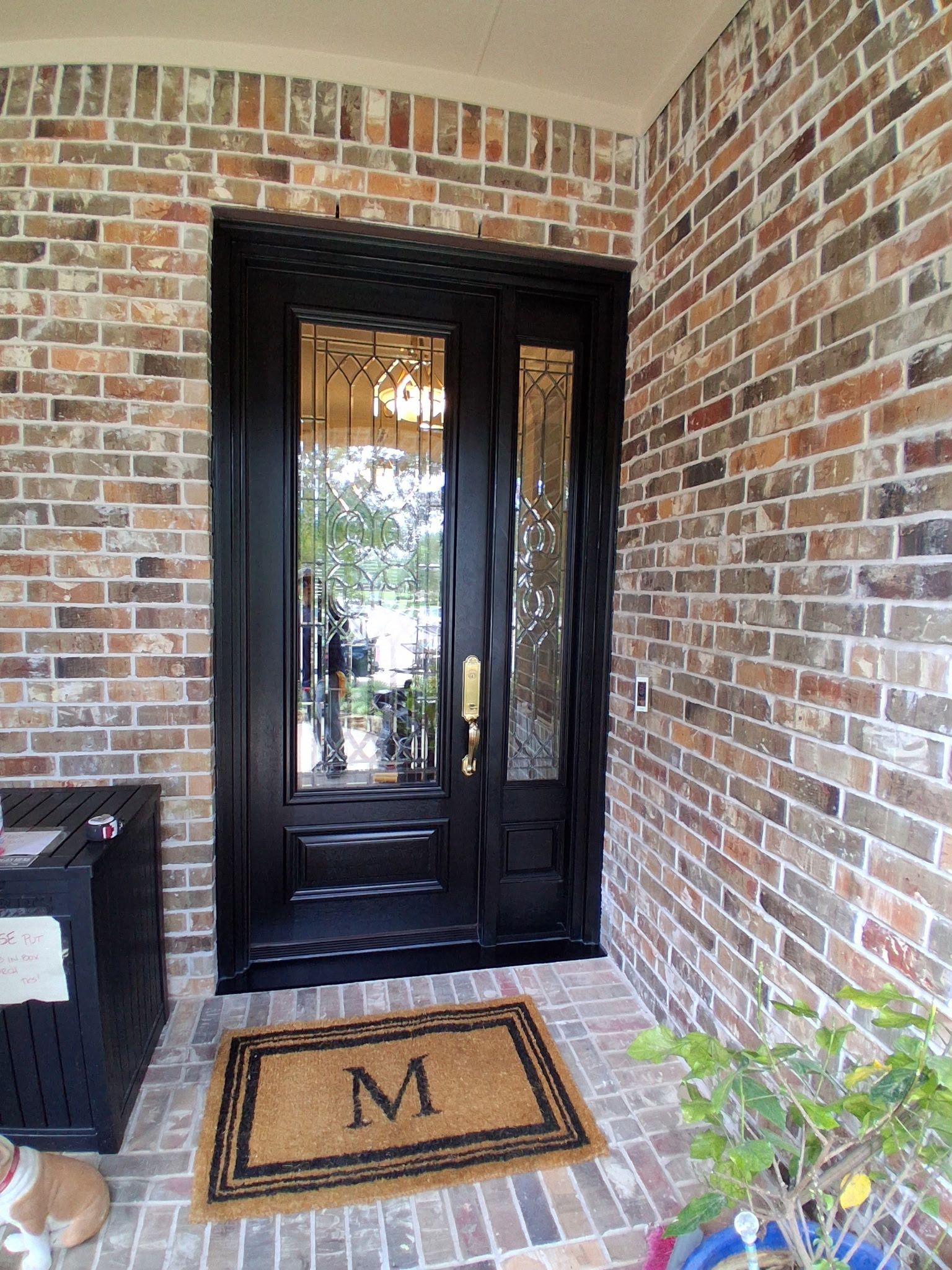 Brick front porch with black double glass doors and a tan doormat with the letter M.
