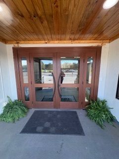 Wooden double glass doors beneath a wood-paneled ceiling, with potted greenery and a black doormat.