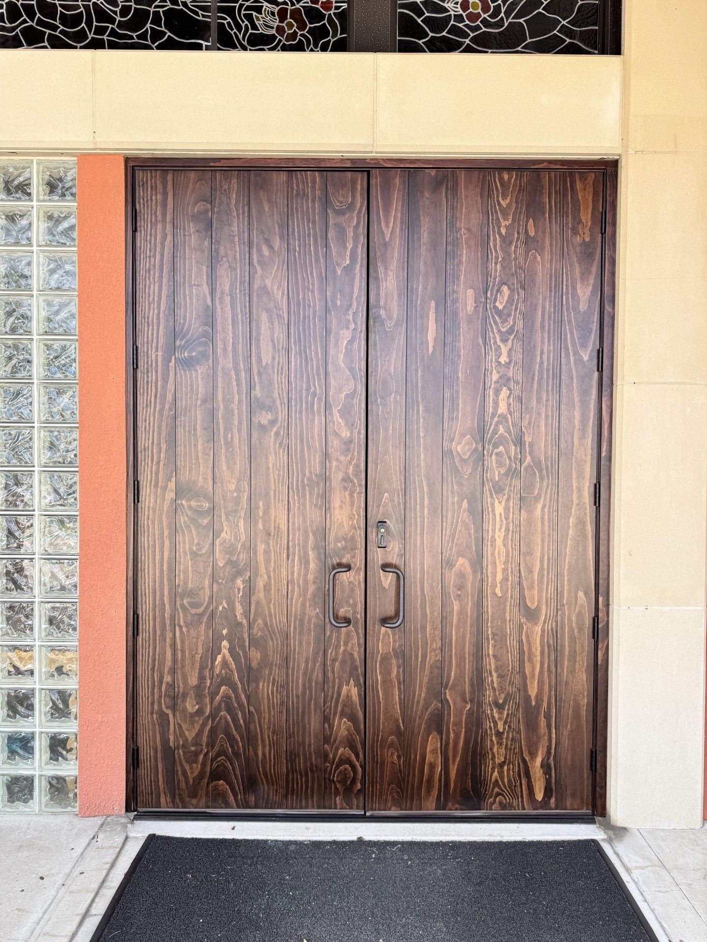 Wooden double doors with black handles set in a beige and glass-block wall.