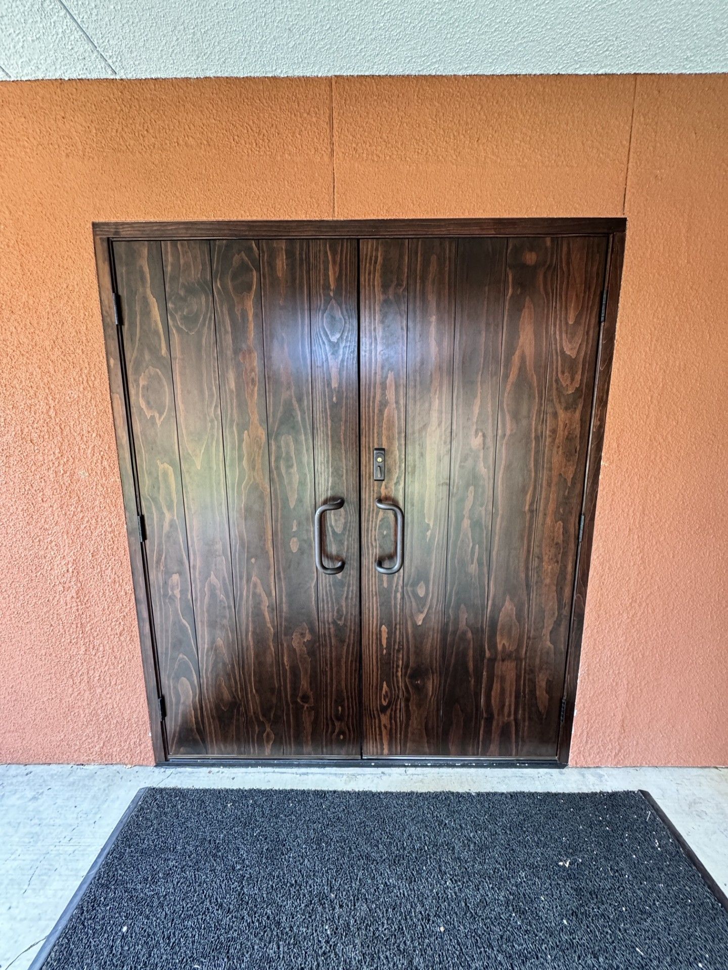 Closed double wooden doors in an orange stucco wall, with a dark floor mat in front.