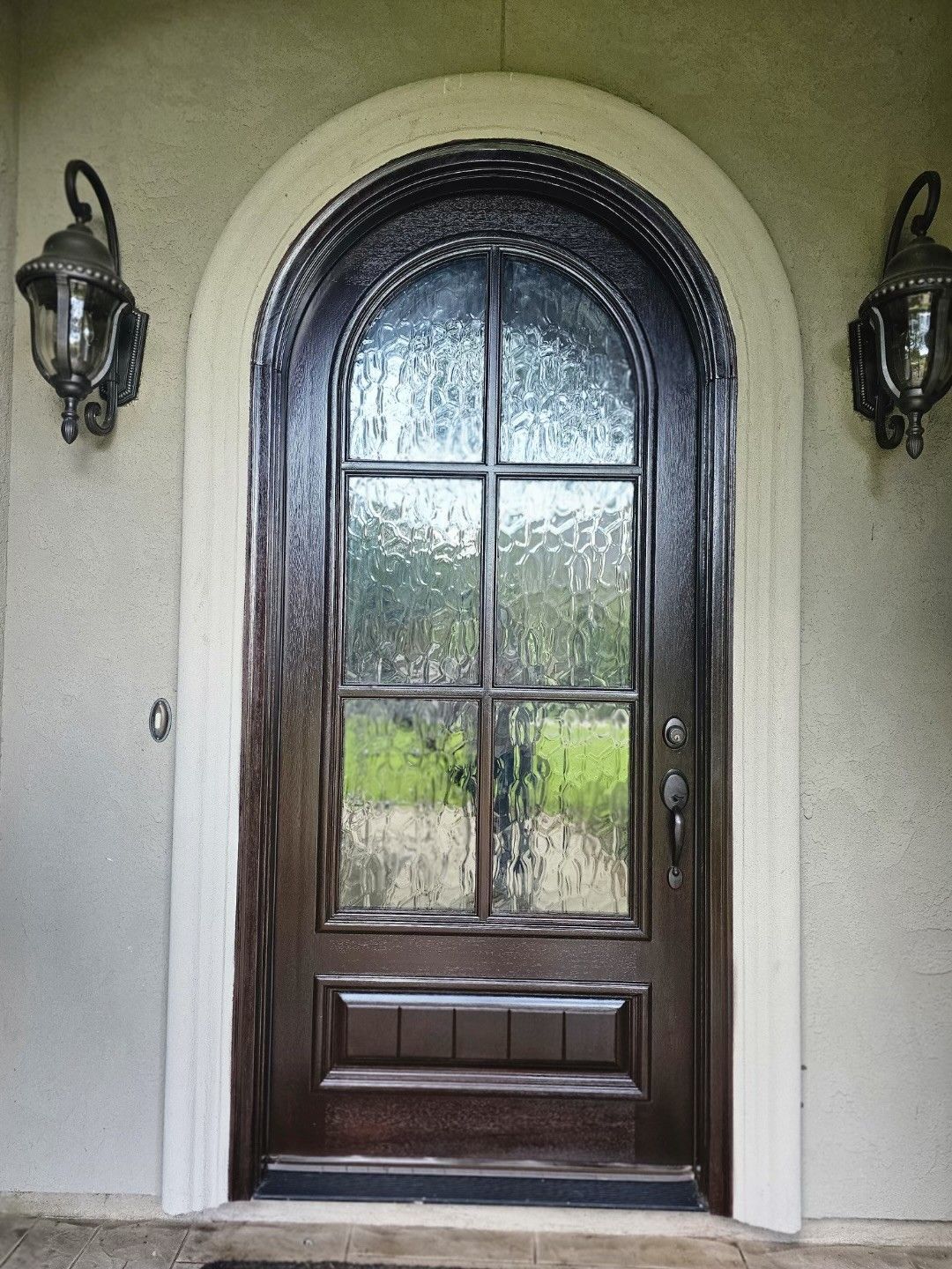 Dark wooden arched front door with glass panels, flanked by two wall lanterns on a stucco entryway.