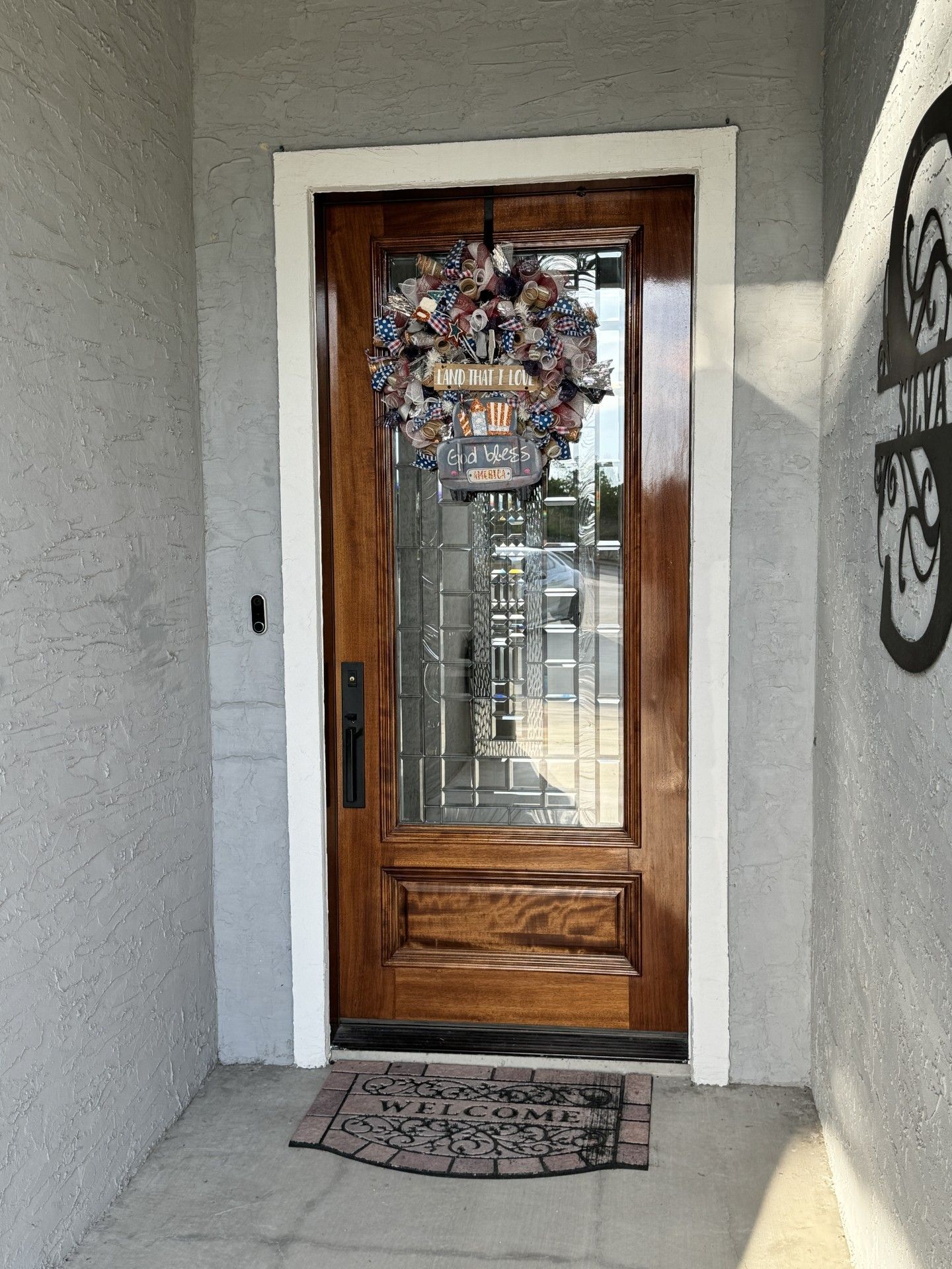 Wooden front door with glass panels, wreath, and doormat in a stucco entryway.