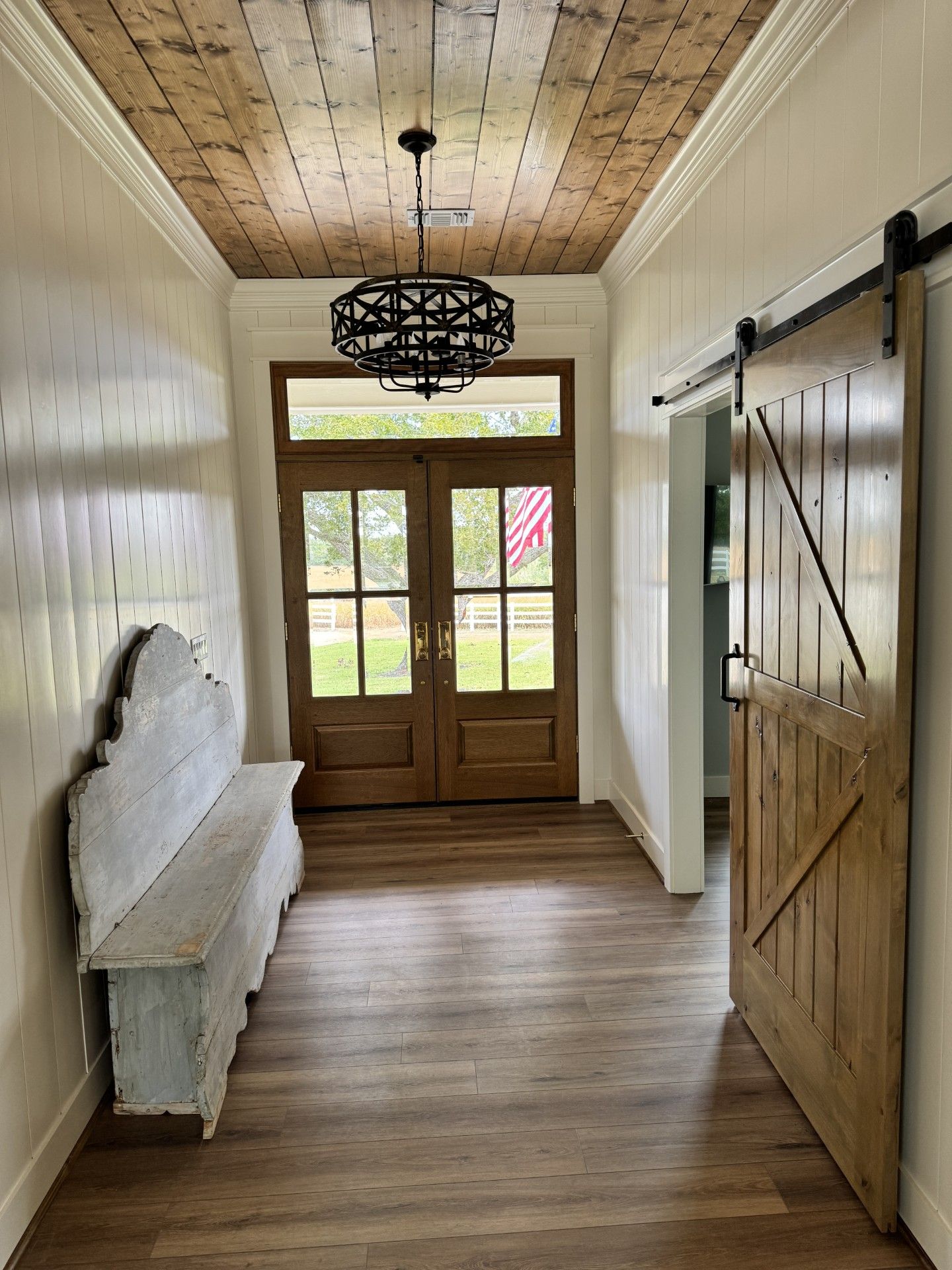 Narrow farmhouse hallway with wood floors, a white bench, barn door, and double front doors.