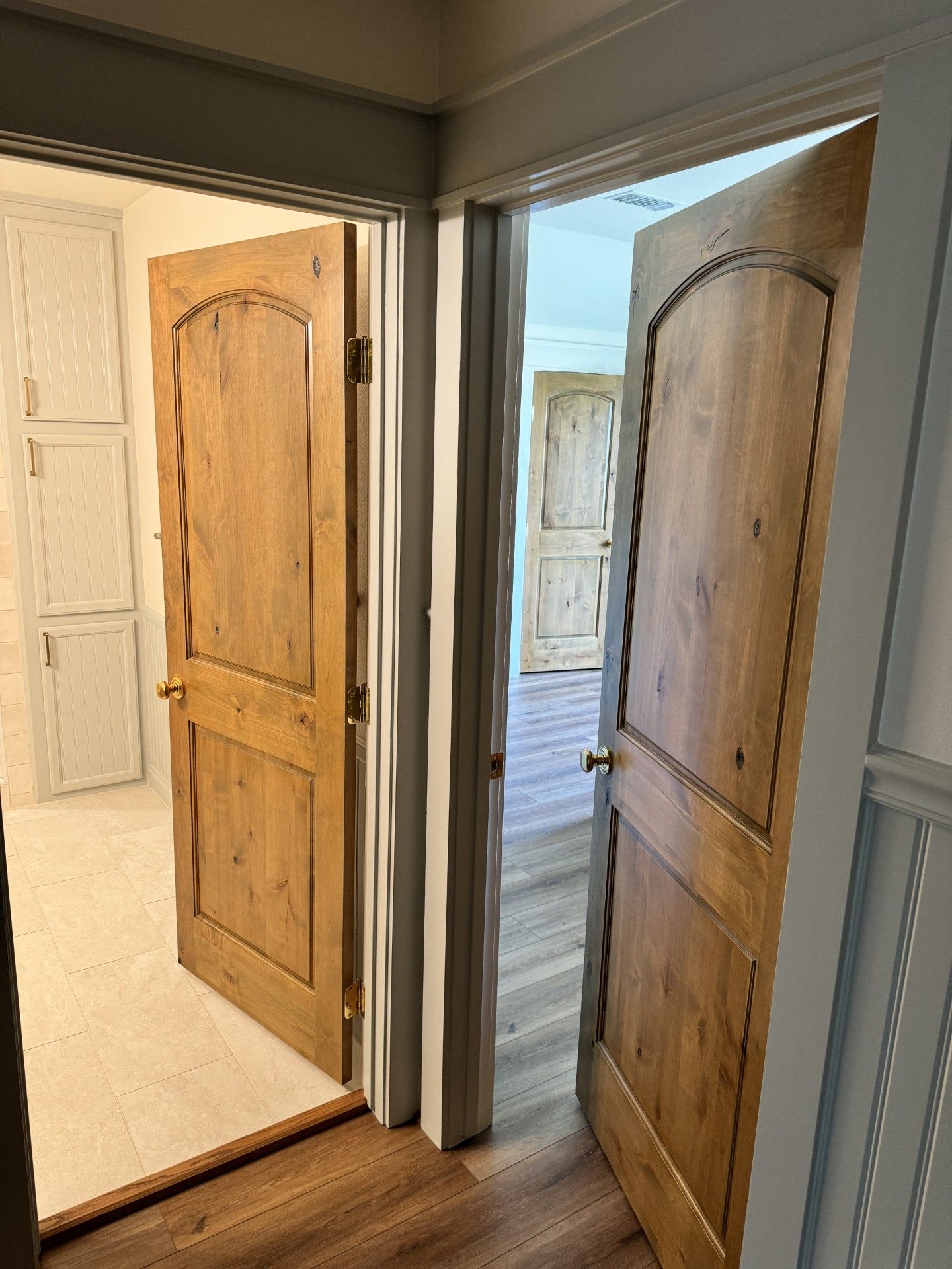 Two open wooden interior doors in a hallway, one leading to a bright room and one to a bedroom.