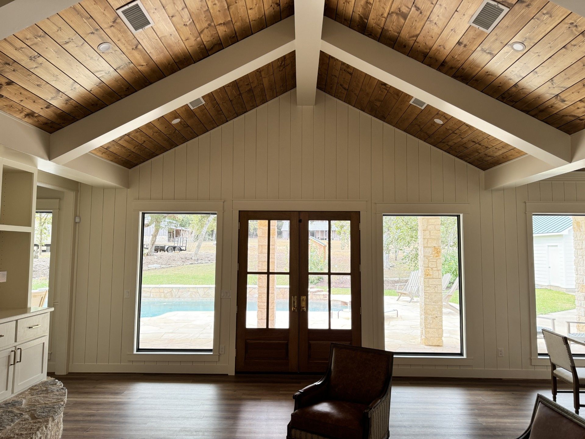 Empty living room with vaulted wood ceiling, French doors, and large windows overlooking a pool.