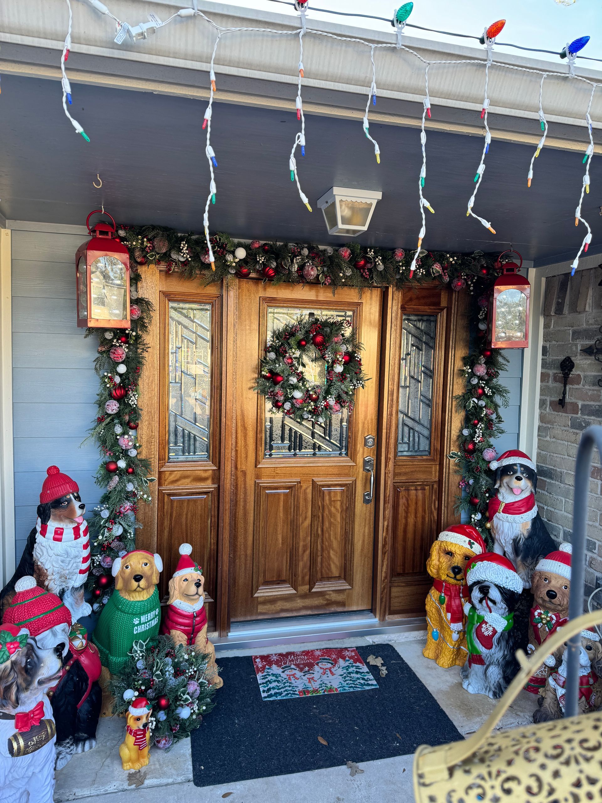 Christmas-decorated front porch with wreaths, lights, garland, and Santa and snowman figures by wooden double doors