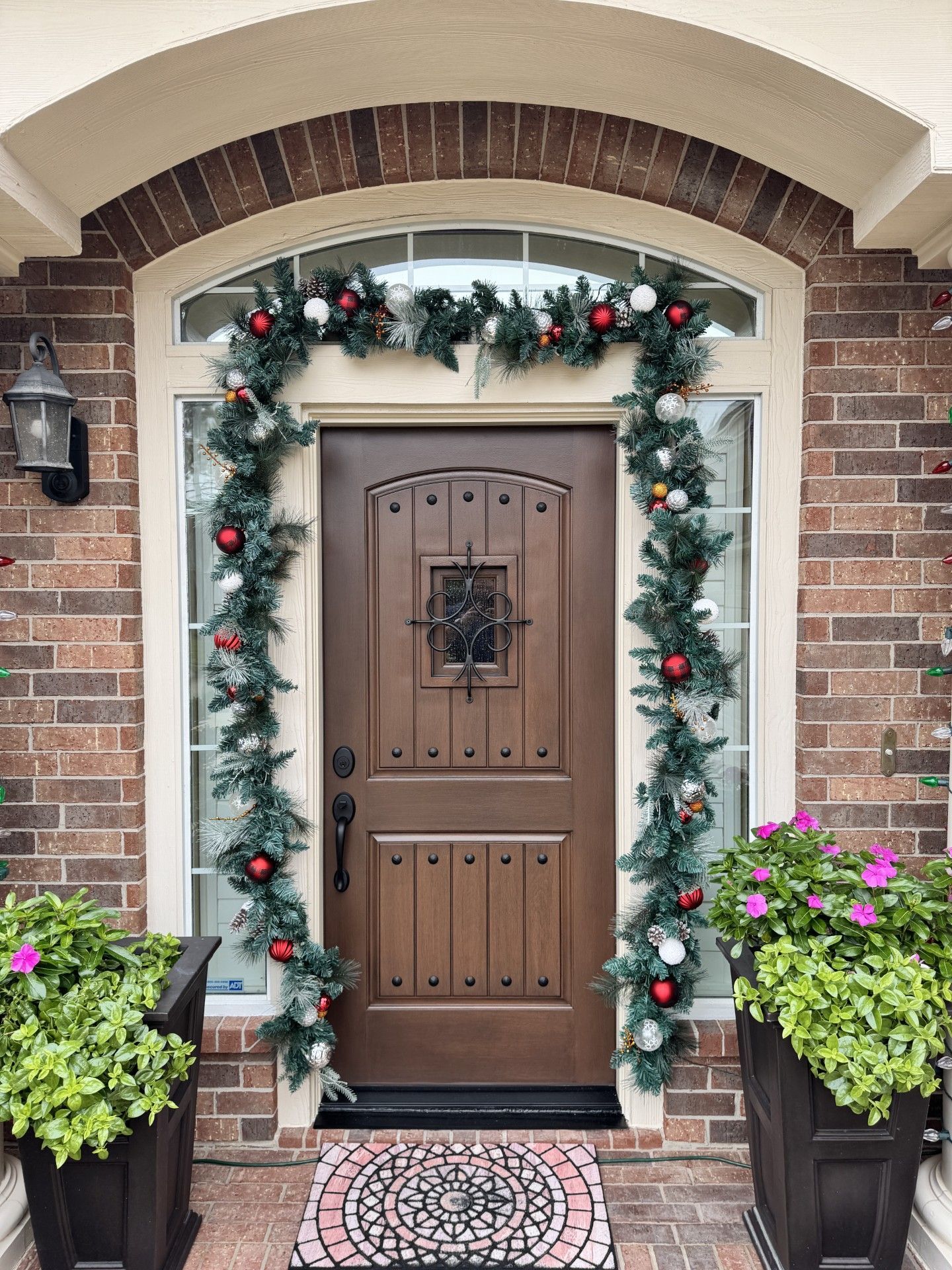 Front porch with a decorated brown front door, brick arch, holiday garland, and potted flowers.