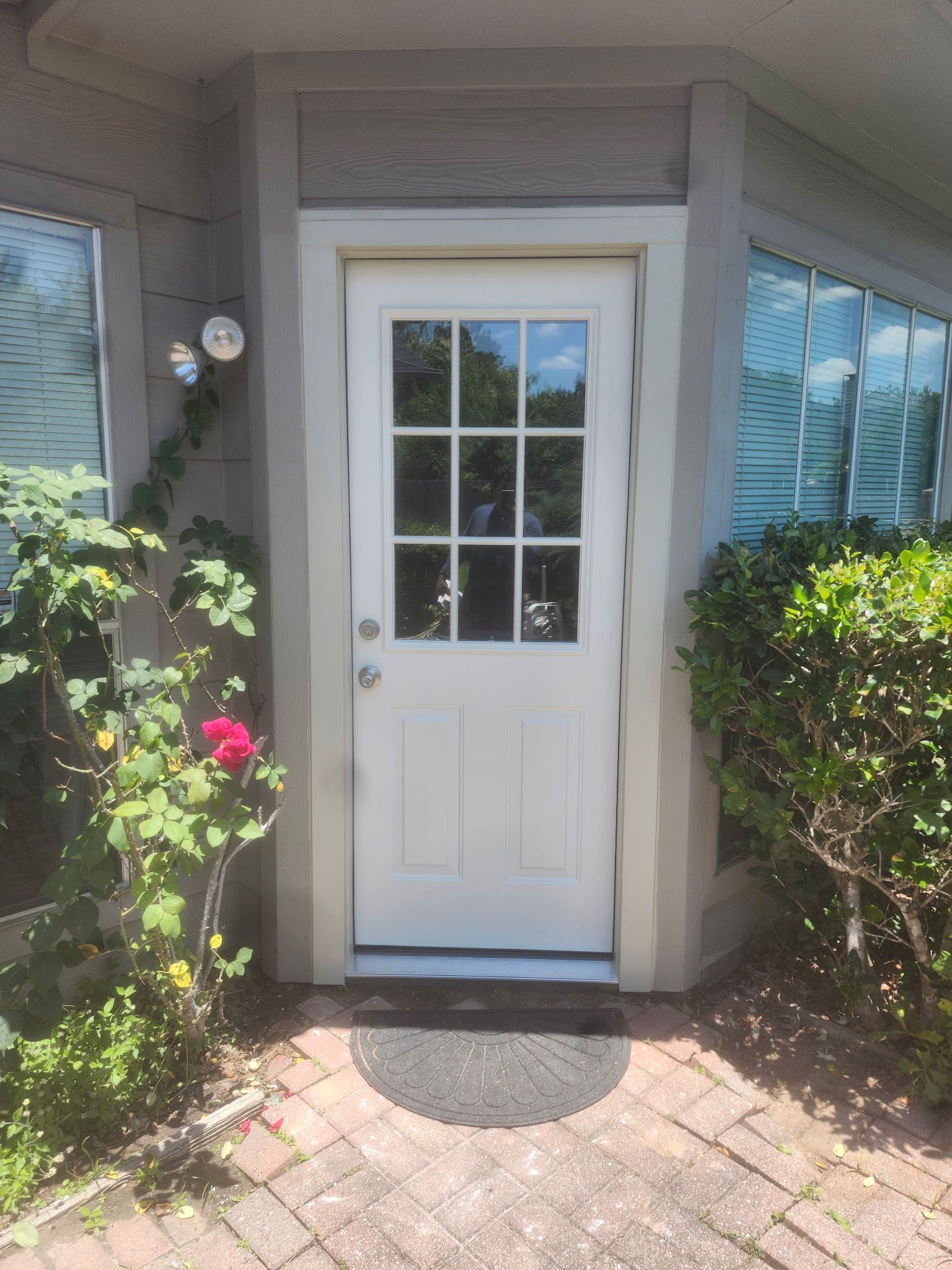 White front door with window panes, centered between bushes and blue shutters, at a brick entryway.
