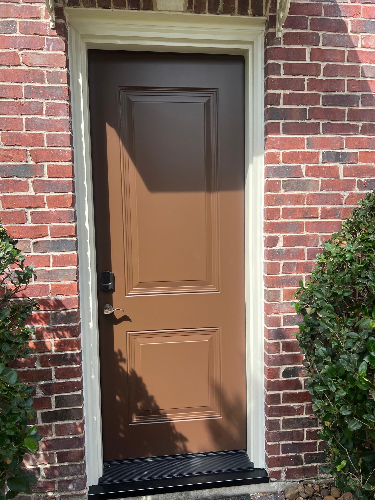 Brown front door with black hardware set in a red brick exterior wall, lit by sunlight.