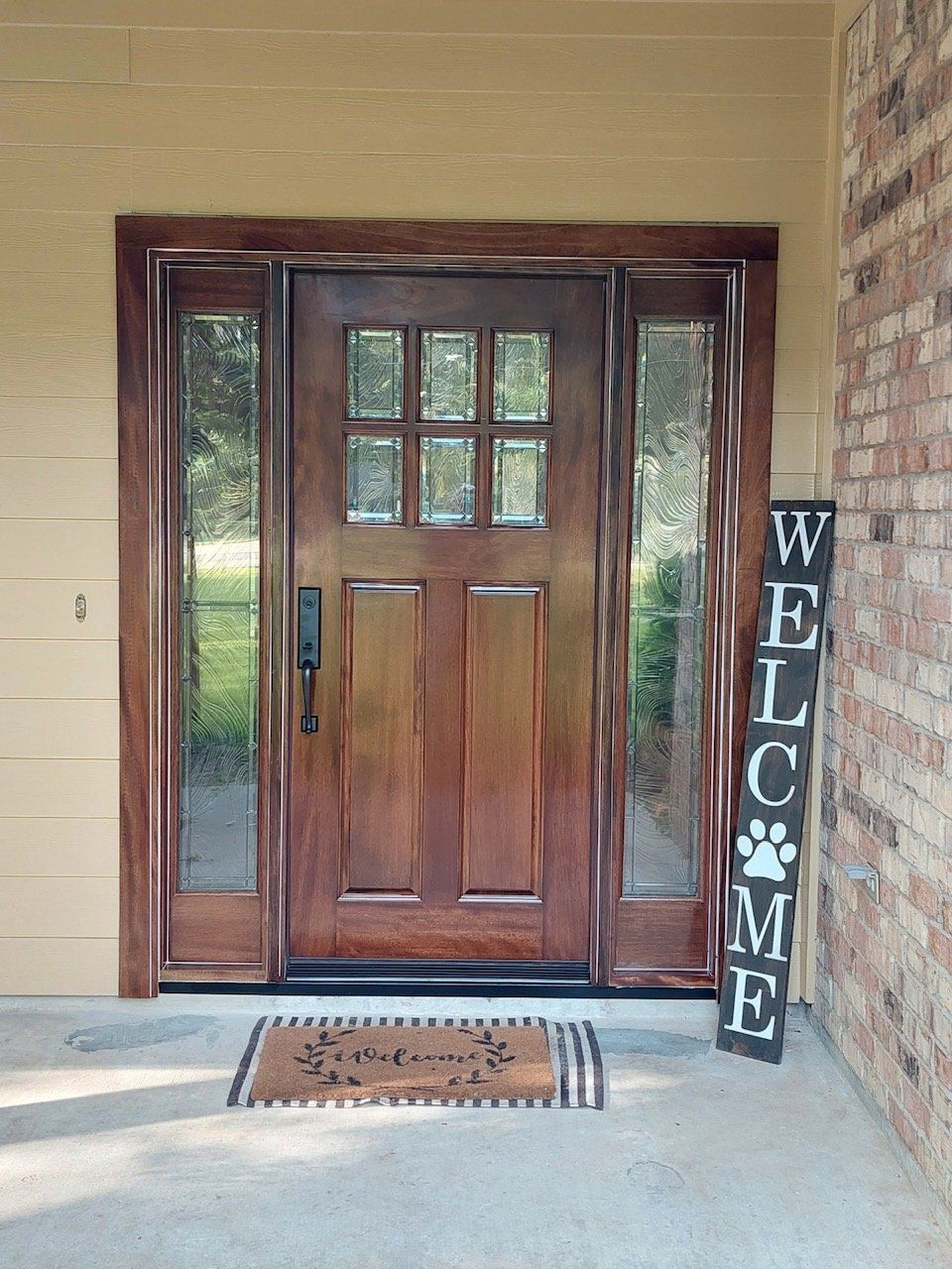 Front porch with wooden front door, side windows, welcome sign, and doormat.