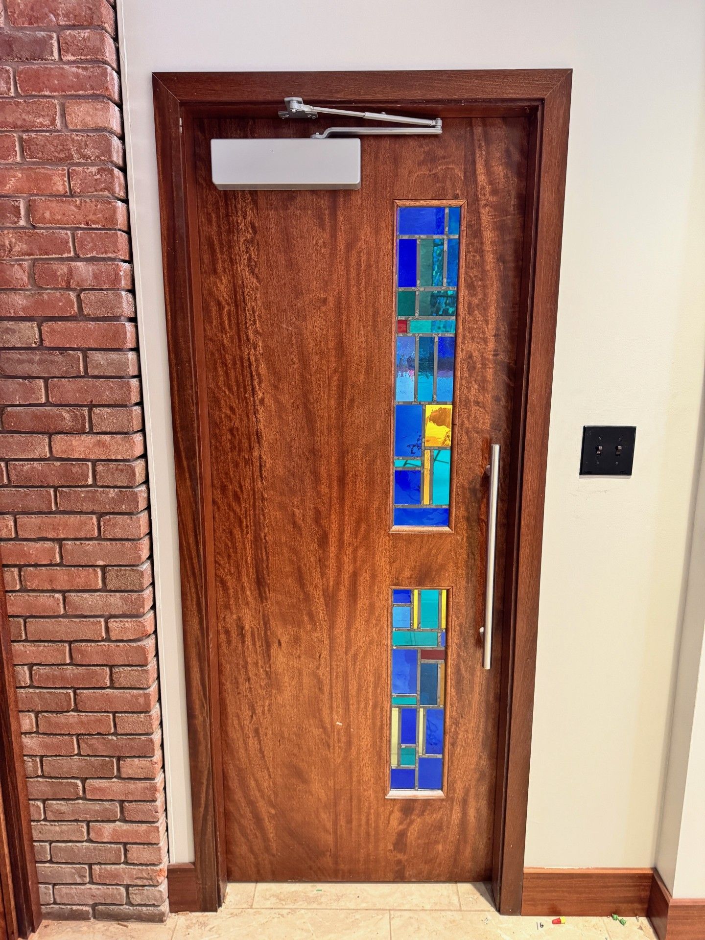 Wooden door with narrow blue stained-glass panels beside a brick wall and cream hallway.