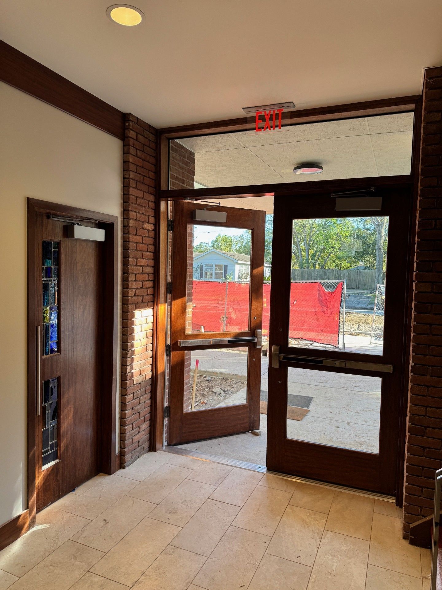 Building lobby with glass exit doors, tiled floor, and sunlight showing red construction fencing outside
