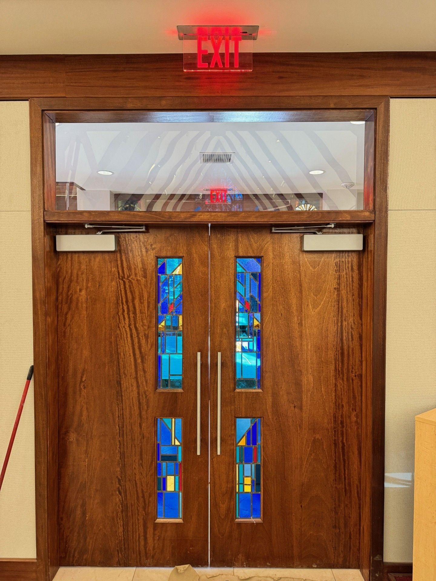 Wooden double doors with blue stained-glass panels beneath a red exit sign.