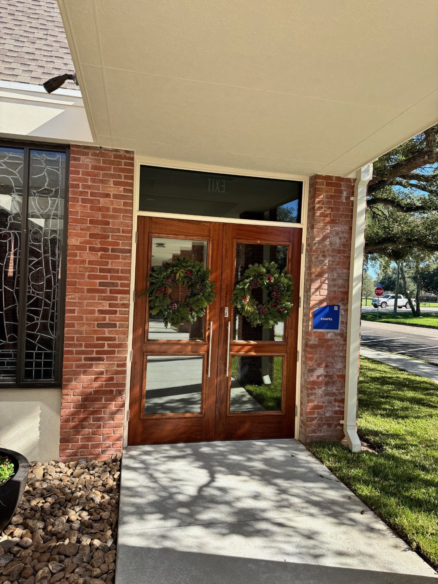 Brick building entrance with wooden double doors, wreaths, and a blue plaque by the walkway