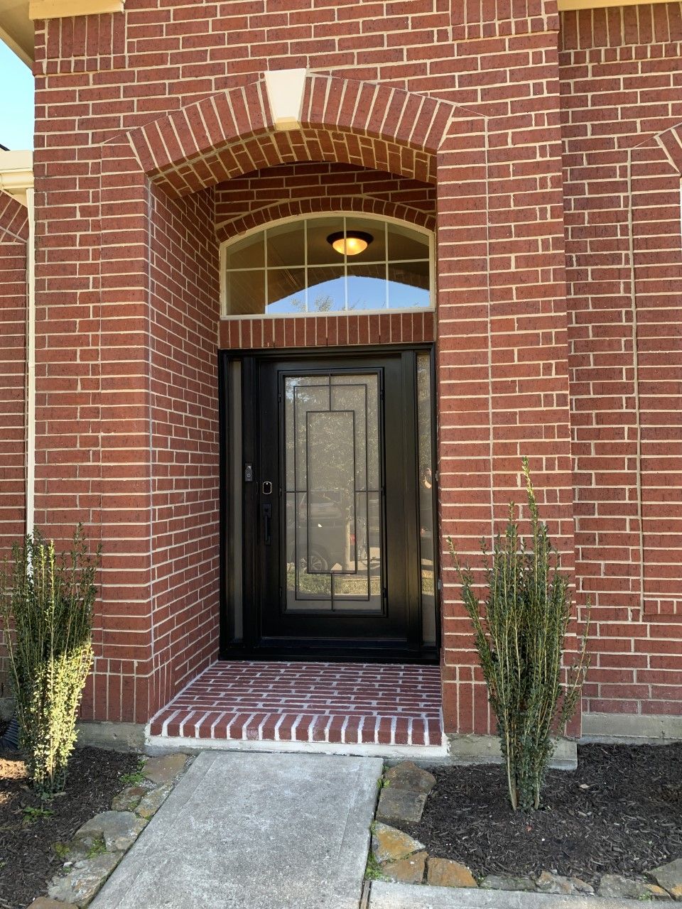 Brick house entrance with black front door, arched brick entry, and potted shrubs on either side