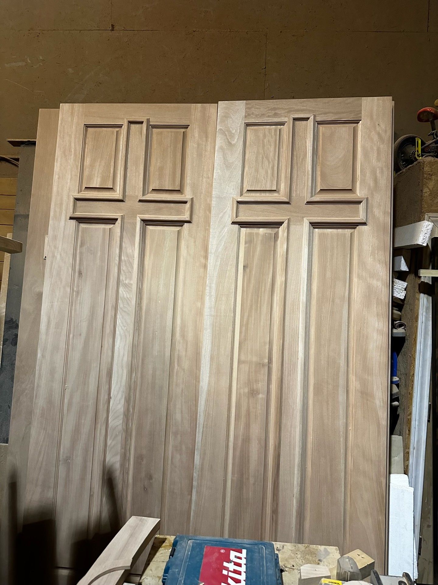 Two unfinished wood cabinet doors standing upright in a workshop, with tools visible at the bottom.