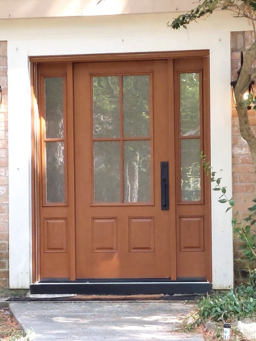 Wooden front door with glass panes and a black handle, framed by white stucco and greenery