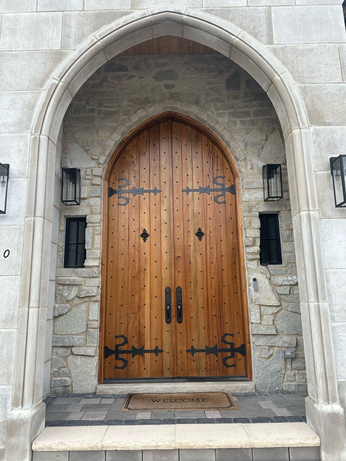 Wooden arched doorway in a stone wall, with black iron hinges and lanterns on either side.