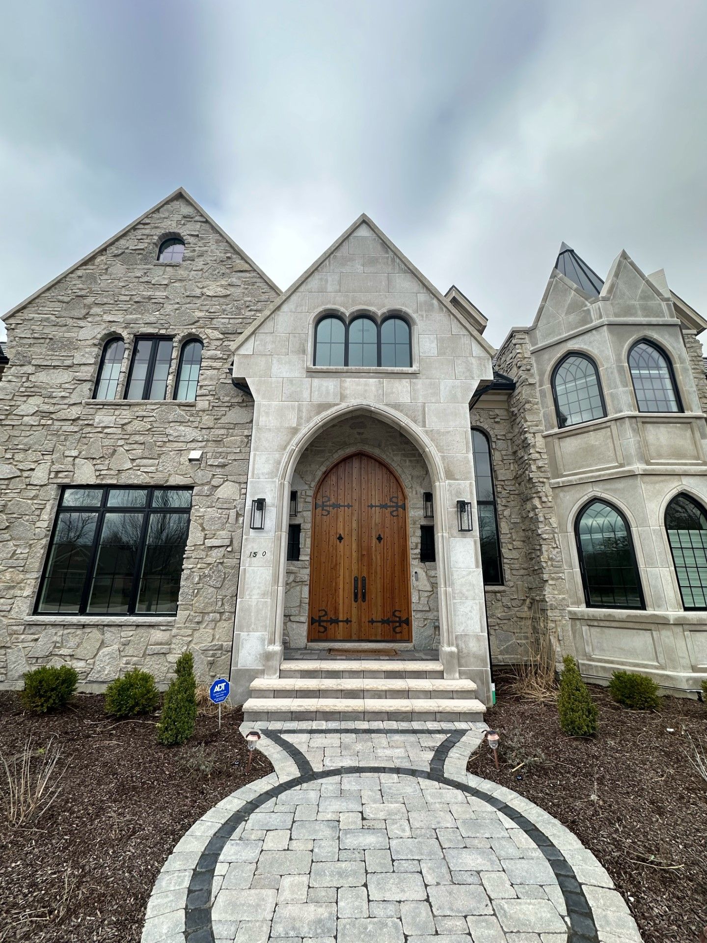Stone house with arched wooden front door, steps, and curved paver walkway under a cloudy sky