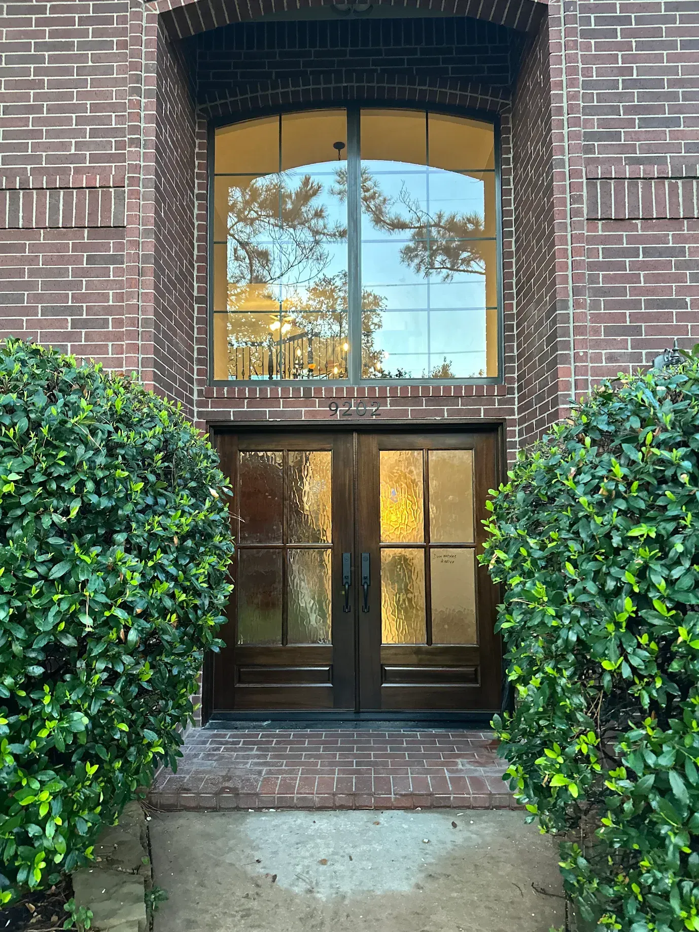 Brick building entrance with glass double doors, arched window above, and trimmed bushes on both sides.