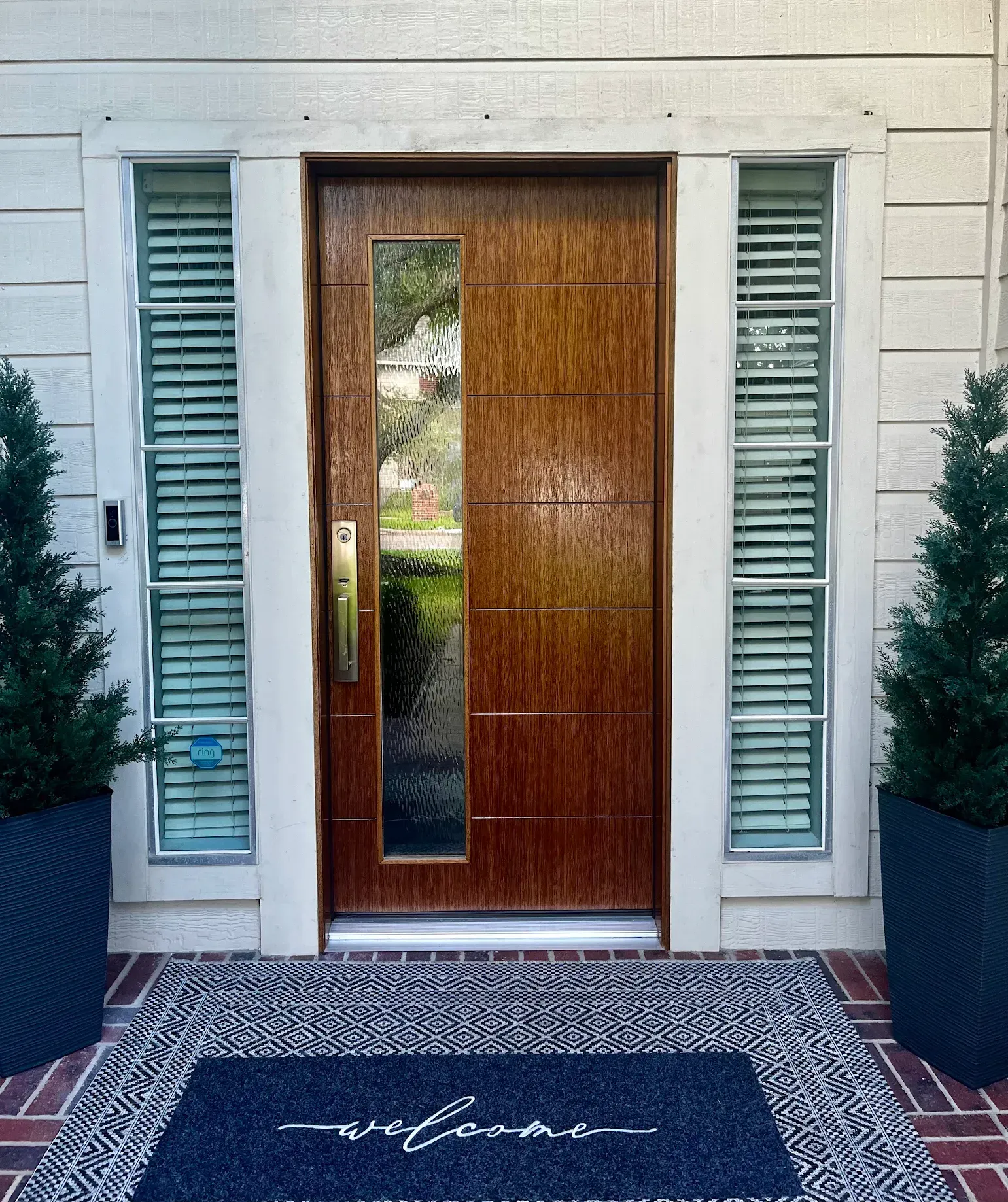 Front door with wood panel, sidelights, and navy “welcome” doormat on a brick entryway.
