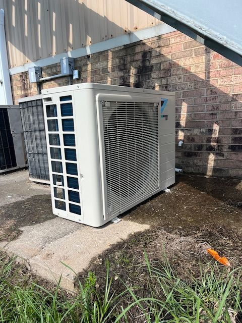 Outdoor air conditioner unit beside a brick wall and metal fence, with grass and wet ground nearby