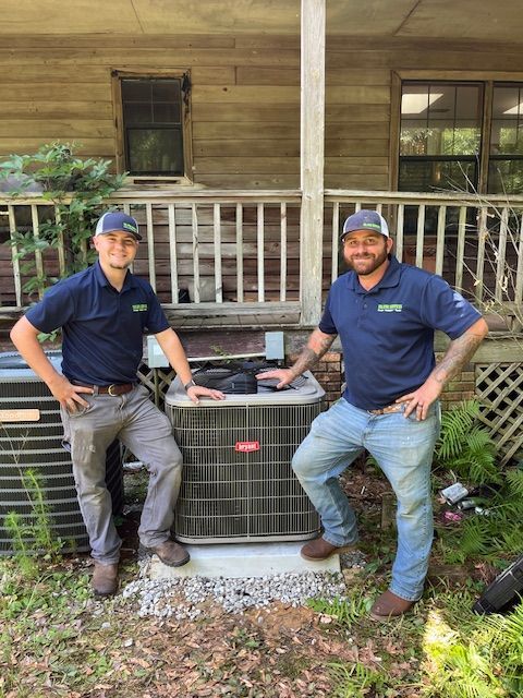 Two workers posing beside a large outdoor HVAC unit in front of a wooden porch