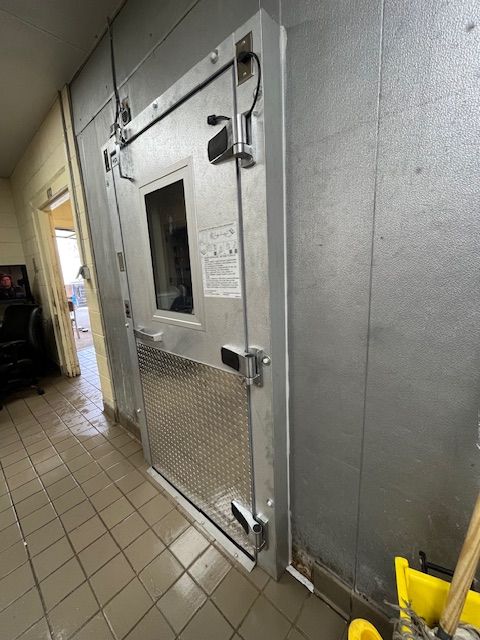 Walk-in industrial freezer door in a tiled hallway with a small window and metal latch.