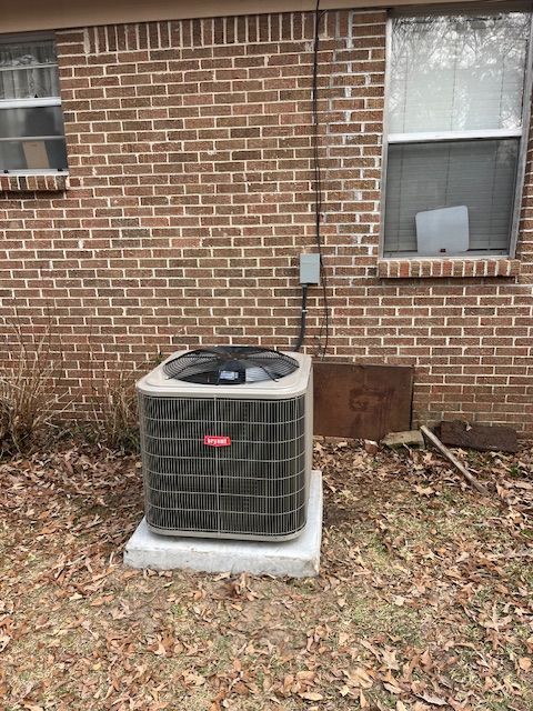 Outdoor air conditioner unit beside a brick house wall with two windows and leaf-covered ground