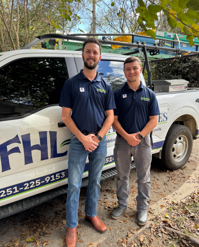 Two men in navy shirts stand beside a white work truck with ladders parked under trees.