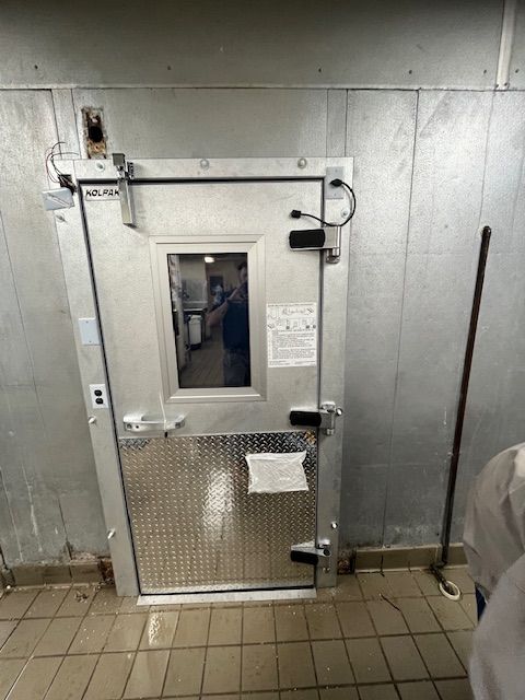 Metal emergency door with a small window and handle in a tiled industrial hallway.