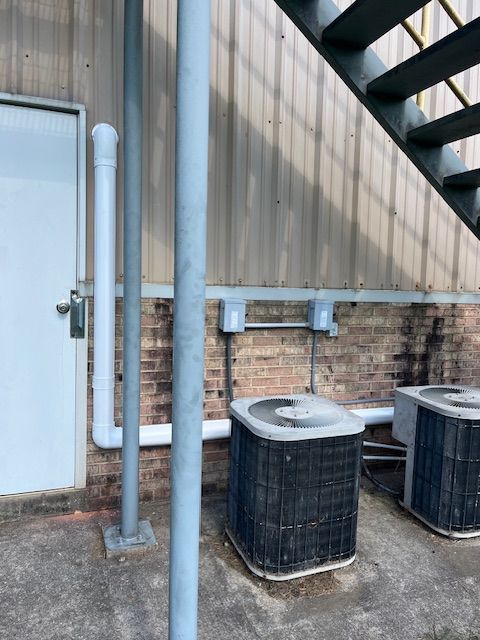 Outdoor HVAC units beside a brick wall under metal stairs, with white pipes and a striped beige siding wall.
