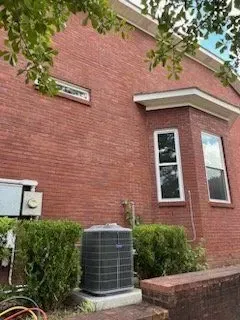 Brick building exterior with HVAC unit, windows, and leafy branches overhead