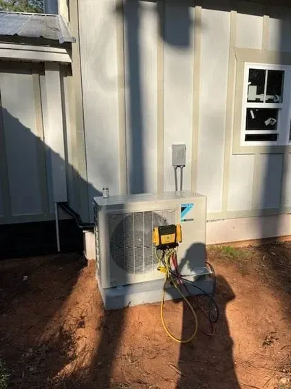 Outdoor HVAC unit beside a house wall, with a yellow tool and cords on the ground