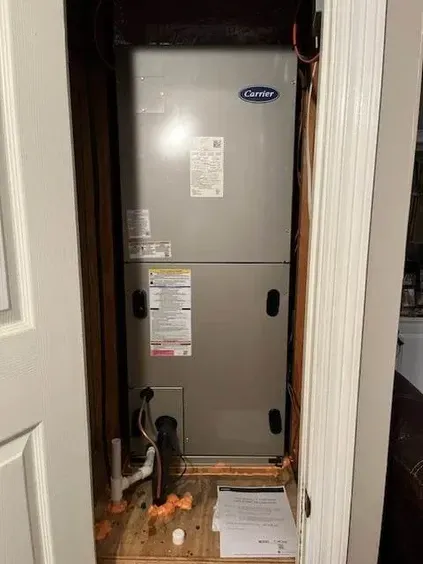 Open furnace in a narrow closet with exposed wiring and pipes, white walls and a door frame.