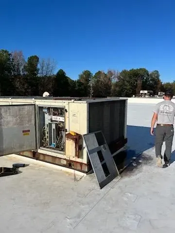 Broken HVAC unit on a rooftop with an open panel and a man walking nearby under a blue sky