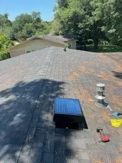 Rooftop view with dark shingles, a solar attic fan, and tree-lined houses in the background
