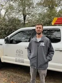 Man standing beside a white service truck in a wooded area, wearing a gray jacket