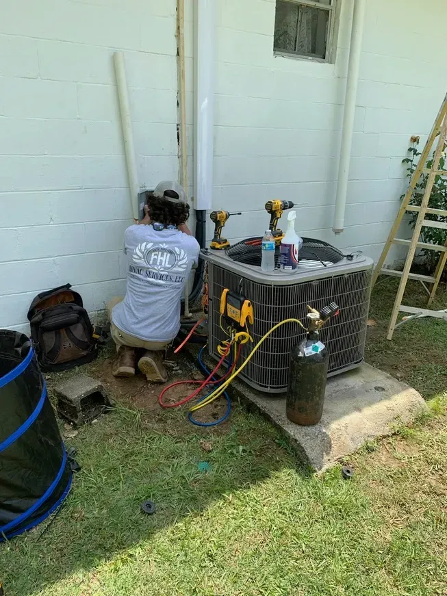 Technician kneels by outdoor HVAC unit, using tools against a white house wall.