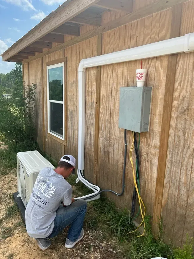 Worker kneeling beside a house, connecting white conduit and wiring to an exterior electrical box.