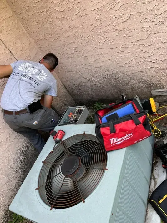Technician working beside a large outdoor HVAC unit with tools on top, against a stucco wall.