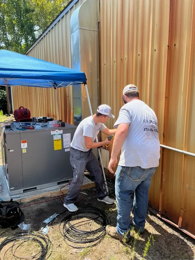 Two workers repairing equipment beside a tan wall, with cables on the ground and a blue canopy nearby