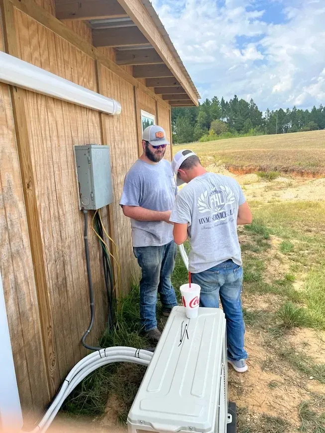 Two workers stand beside a wooden shed, handling a white utility unit and hoses outdoors.