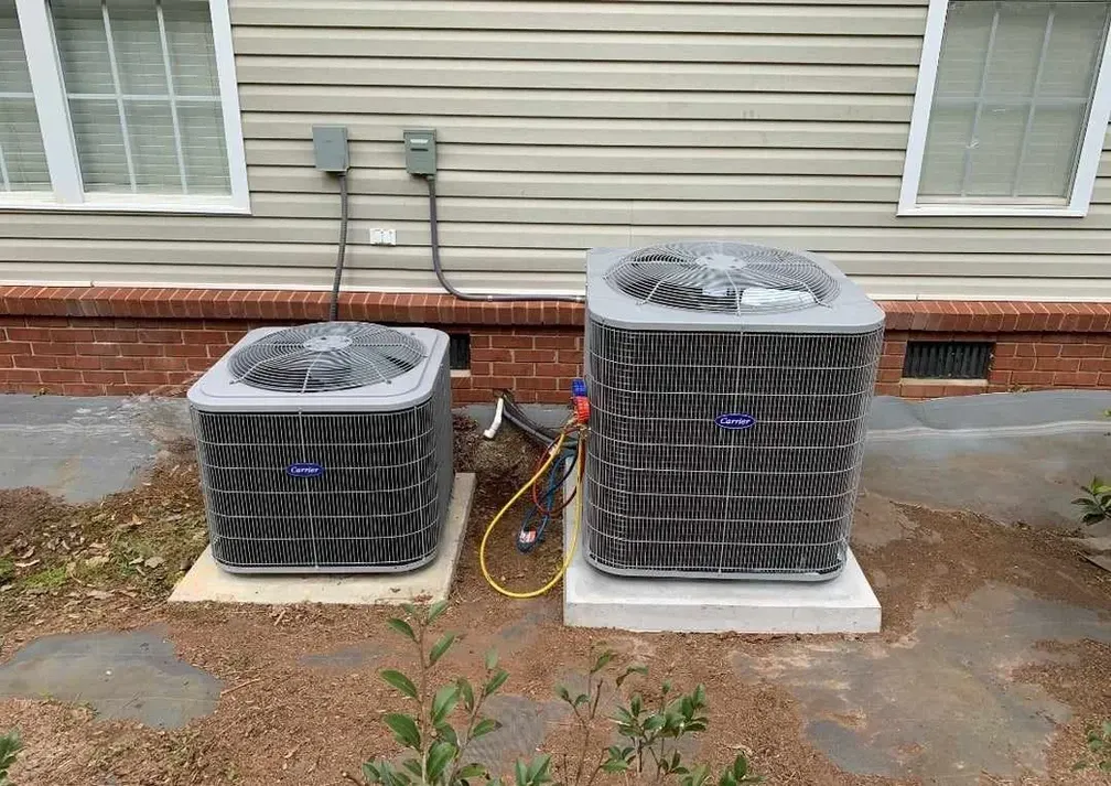 Two outdoor air conditioning units beside a house with beige siding and brick foundation.