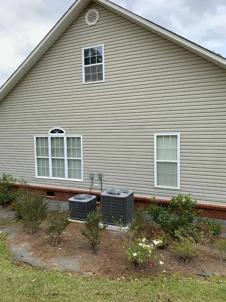 Suburban house side wall with beige siding, two windows, shrubs, and outdoor HVAC units.