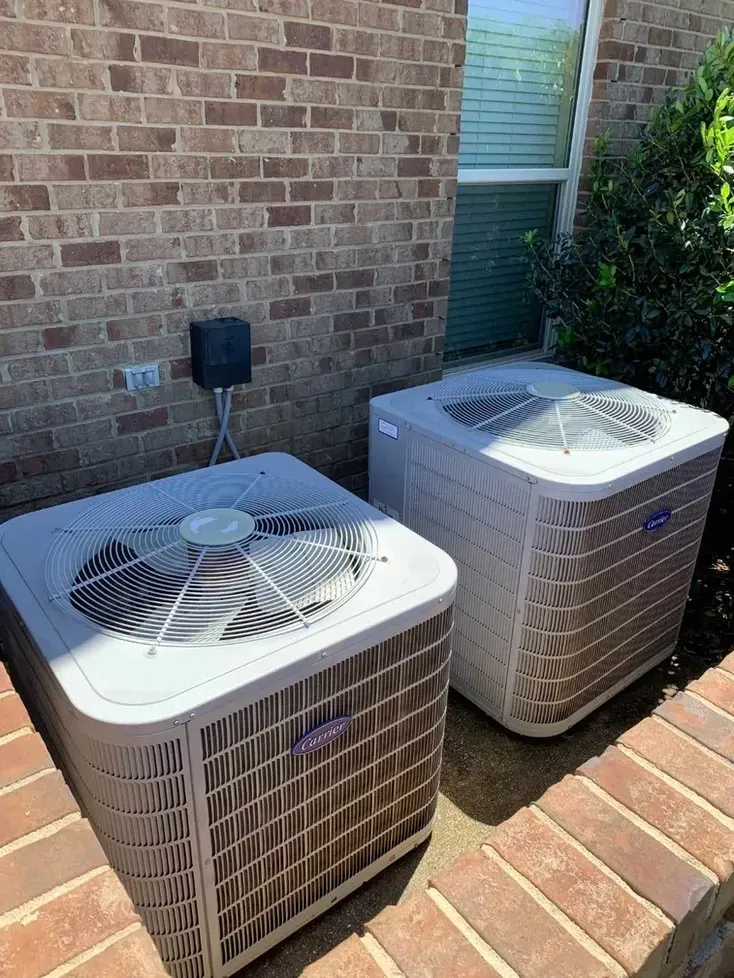 Two outdoor HVAC units beside a brick house wall on a brick patio.